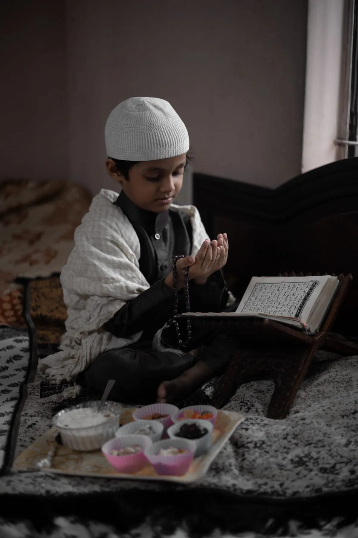 A boy in a white cap raising his hands in supplication beside an open Quran