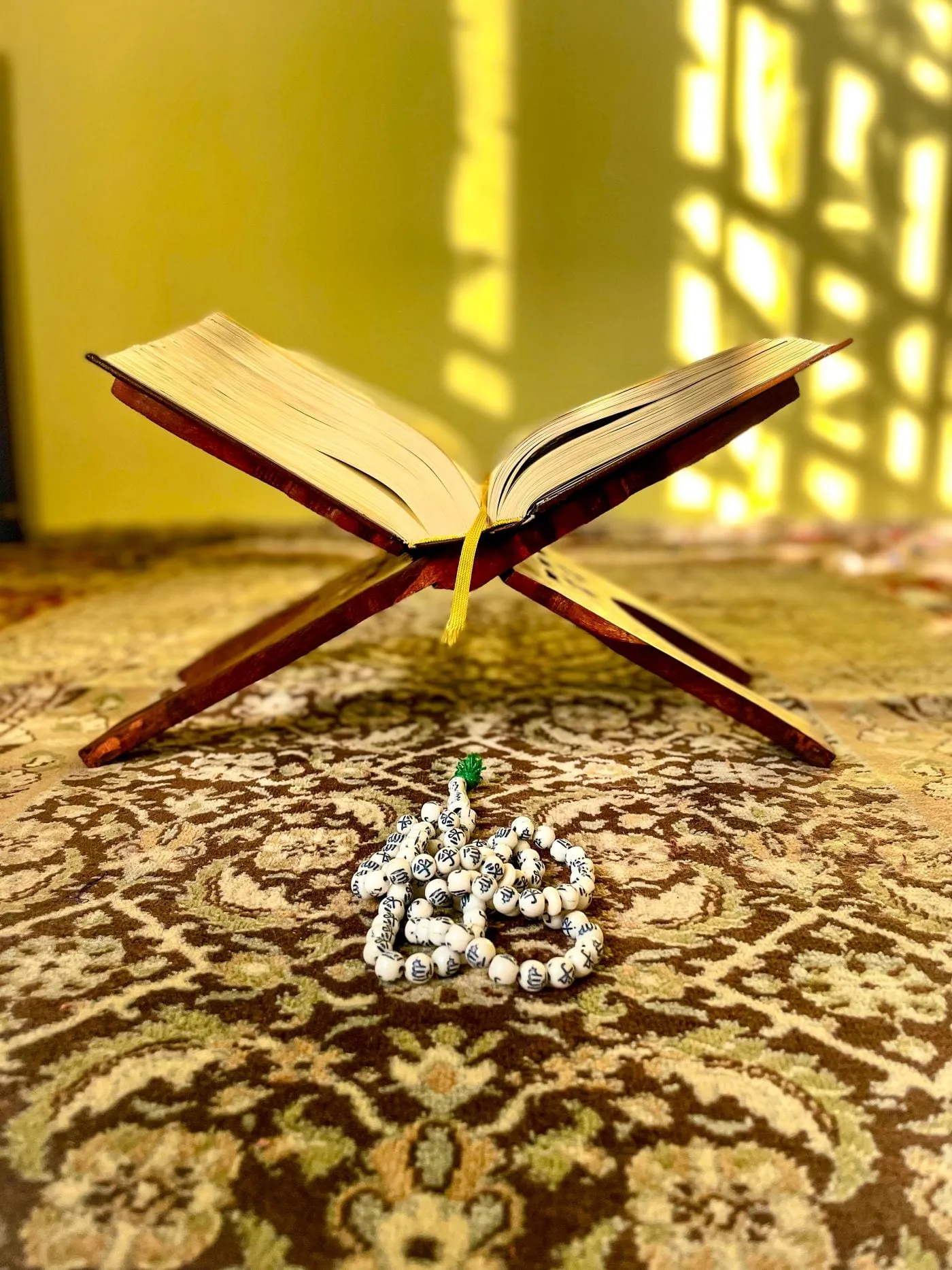 An open Quran on a wooden rehal in warm sunlight, with prayer beads beside it