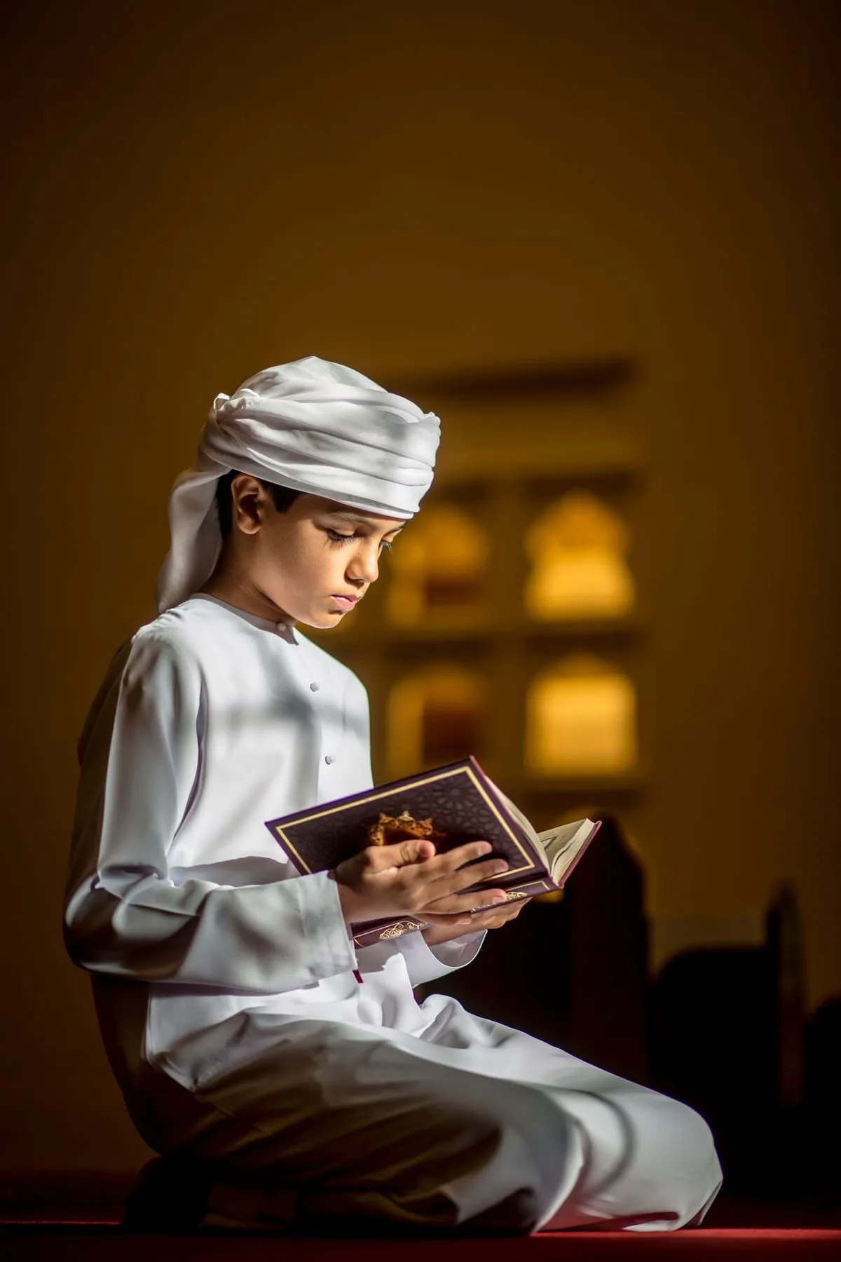 A boy in white reading the Quran in warm mosque light