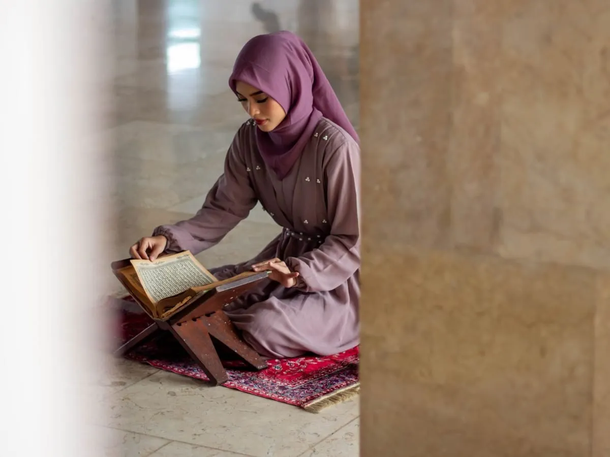 A woman reading the Quran on a stand while seated on a prayer mat