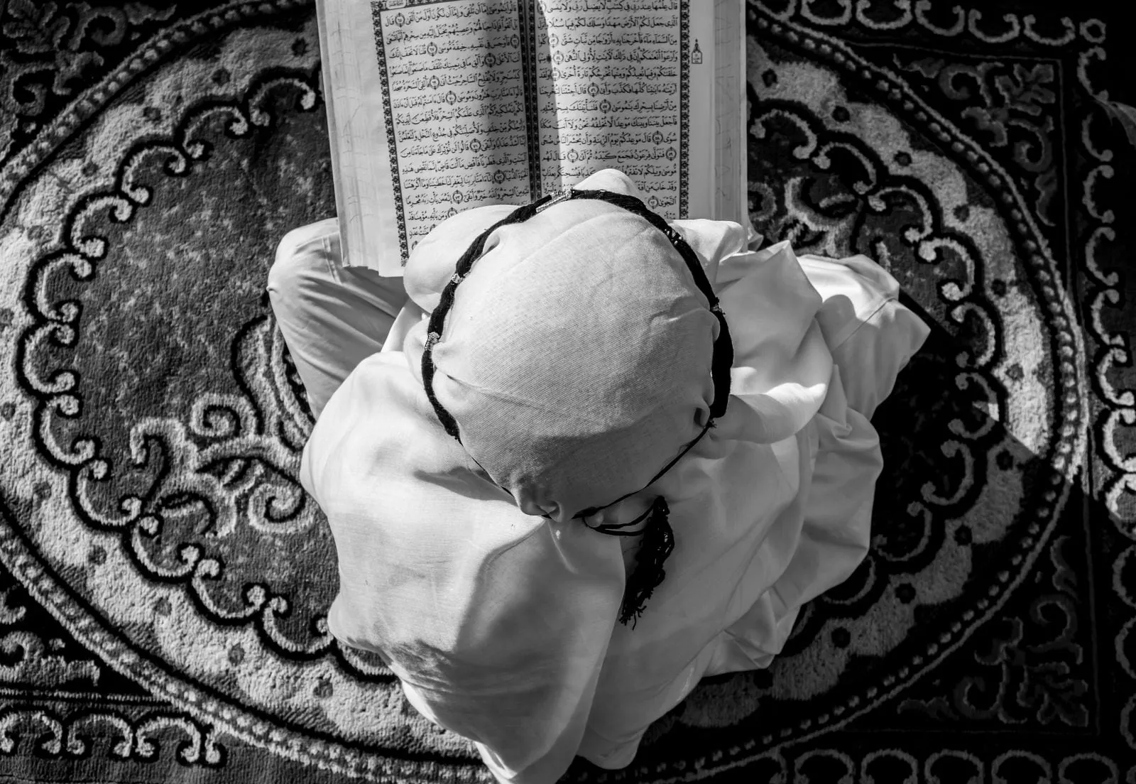Seen from above, a person in white reading the Quran on a patterned rug